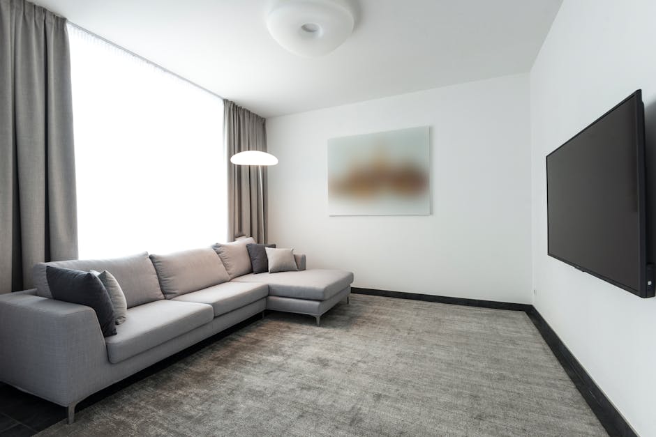 A modern bedroom showing a large flat-screen television mounted on a white wall adjacent to a white sliding door. The room features a bed with a beige upholstered headboard and a white mattress positi