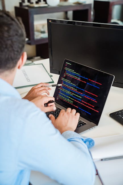 A close-up image of a person's hand using a trackpad on a silver laptop placed on a wooden desk. The laptop screen displays lines of code with syntax highlighting in various colors. The individual is 